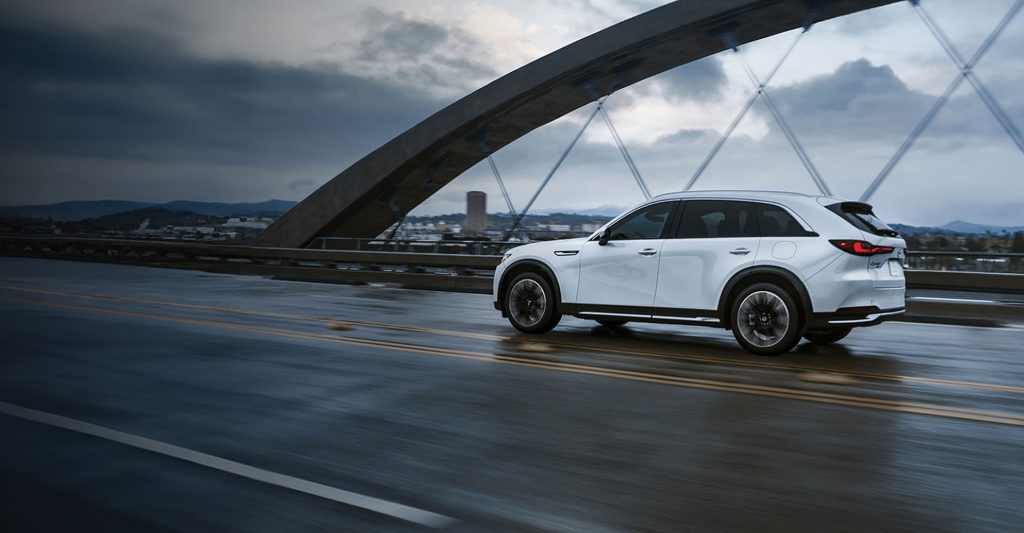 A white Mazda CX-90 PHEV driving across a bridge on a rainy day