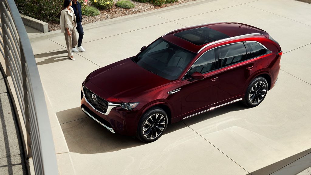 Red 2026 Mazda CX-90 with panoramic sunroof parked on a driveway, couple walking nearby on a sunny day.
