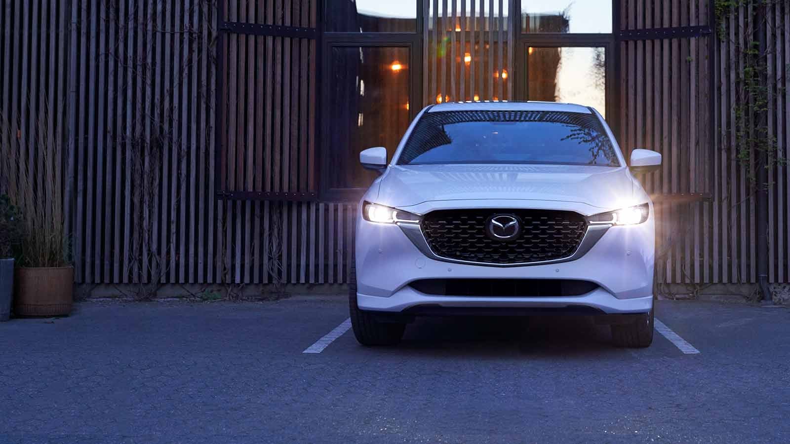 A white Mazda CX-5 parked in front of a modern building with wooden slats, illuminated by its headlights.