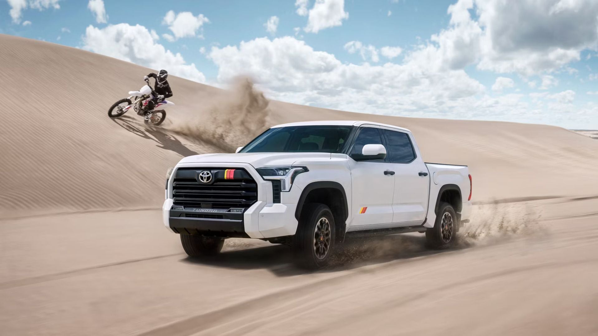 White Toyota Tundra driving on sand dunes beside a motorcyclist kicking up dust.